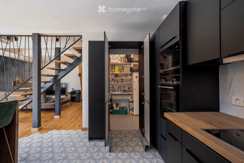 Modern kitchen with dark cabinets, stainless steel appliances, and a wooden island. Adjacent living area features a sofa, armchair, and a staircase leading to an upper level.