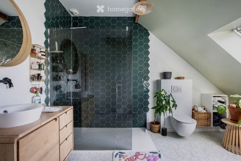 Modern bathroom with hexagonal dark green tiles, a white sink, a shower area, a toilet, and a potted plant. Features include a round mirror, a wooden cabinet, and a small stool with a plant on it.