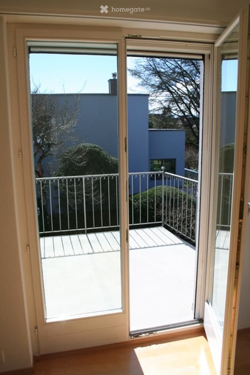Sliding glass doors open to a small balcony with metal railing, overlooking a blue building and garden area.