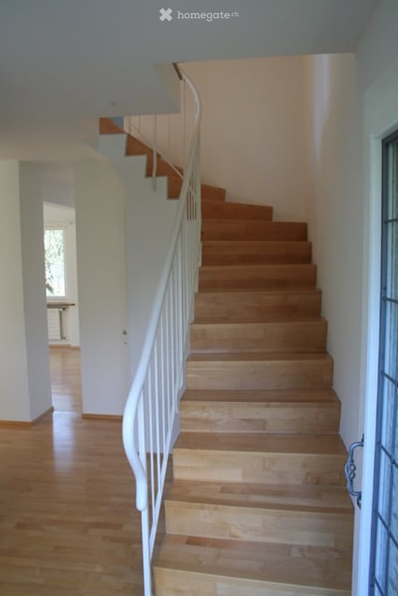 Spacious hallway with wooden flooring and a curved staircase featuring a white handrail. The walls are painted white, and the area is well-lit with natural light coming from an adjacent room.