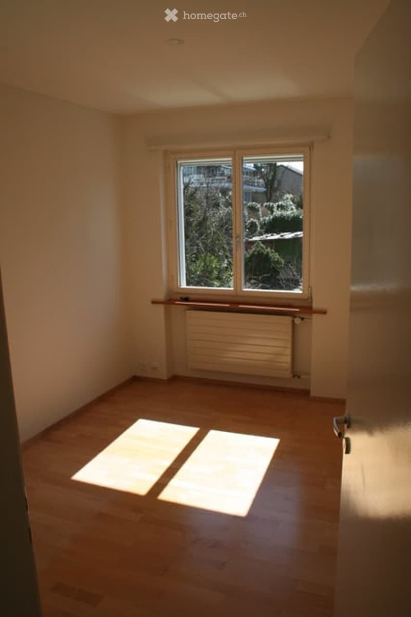 Empty room with wooden flooring, white walls, and a large window with a view of greenery outside. There is a radiator below the window and a small shelf above it.