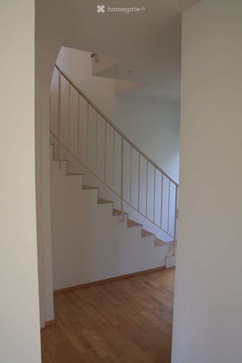 Staircase with wooden steps and white railings, leading to an upper level. Light-colored walls and wooden flooring.