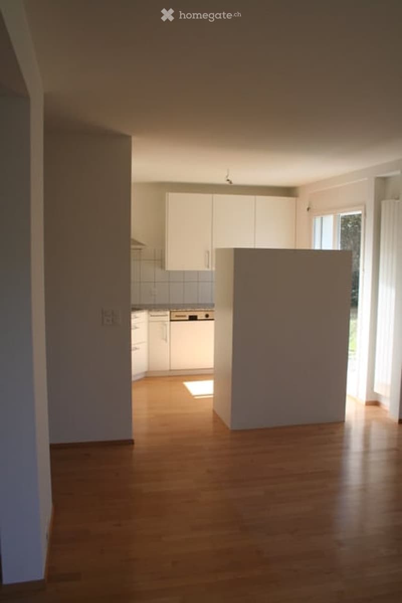 Empty room with wooden flooring, white walls, and a small kitchen area with white cabinets and appliances visible in the background.
