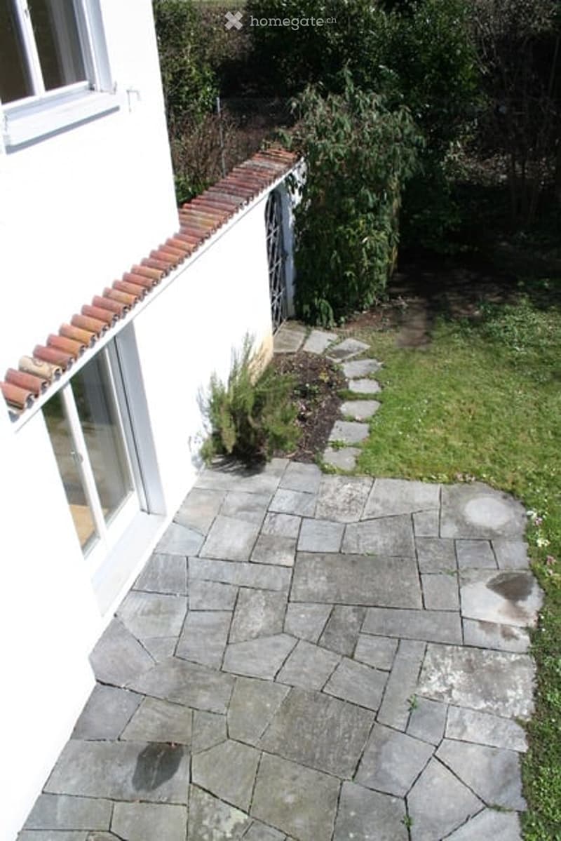 A paved patio area adjacent to a white house with a red-tiled roof. The patio is made of large, irregularly shaped stone tiles and is bordered by a low stone wall. There is a small garden bed with plants and shrubs near the house, and the surrounding area