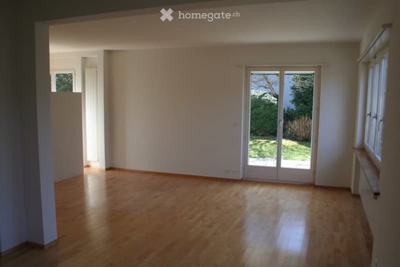 Empty room with wooden flooring, white walls, large sliding glass door leading to a garden, and a window on the right side.