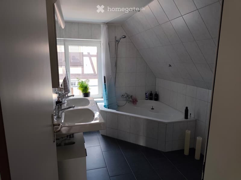 Modern bathroom with a double sink, bathtub, and large window with a view. Includes a plant on the windowsill and candles on the floor.
