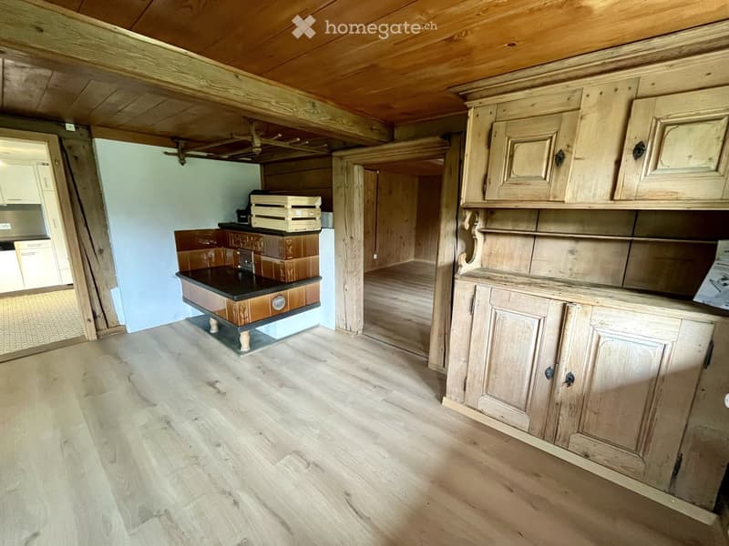 Empty room with wooden flooring, exposed wooden beams on the ceiling, and built-in wooden cabinets. A small black countertop with a sink is visible in the background.