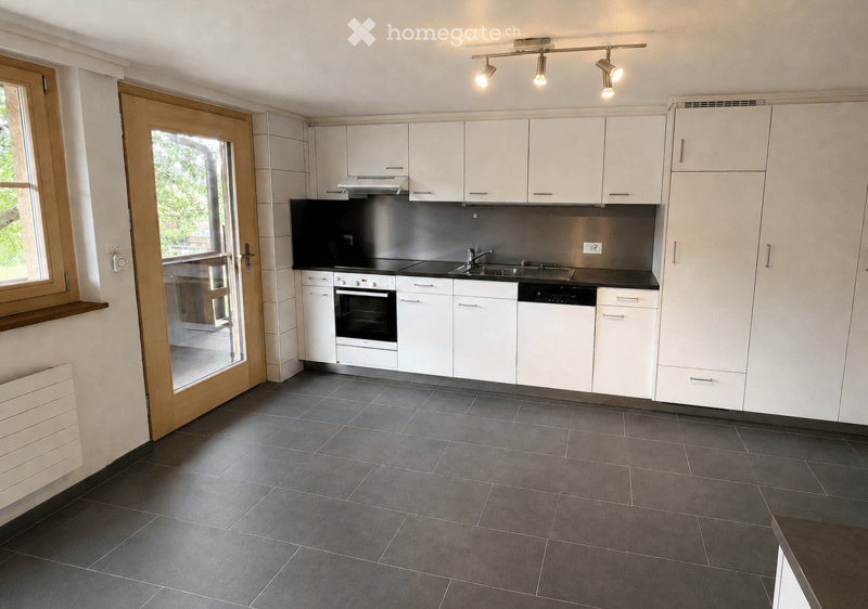 Modern kitchen with white cabinets, dark countertops, and built-in appliances including an oven, sink, and dishwasher. Features track lighting and a large window with a wooden frame.
