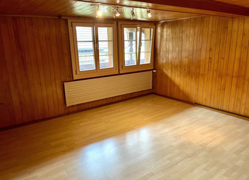 Empty room with wooden walls and floor, large windows, radiator, and ceiling lights.