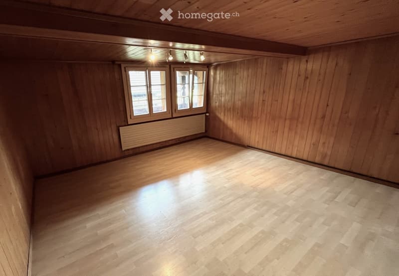 Empty room with wooden walls, wooden floor, and two windows with white frames. A radiator is visible below the windows, and recessed ceiling lights provide illumination.