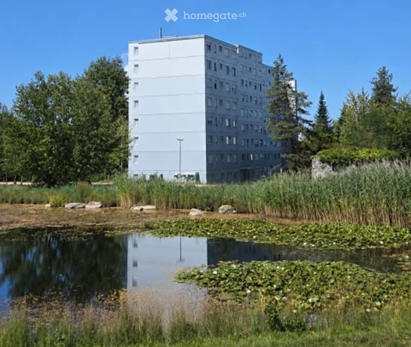 Modern multi-story residential building with white facade and numerous windows, surrounded by greenery and a small pond with lily pads.