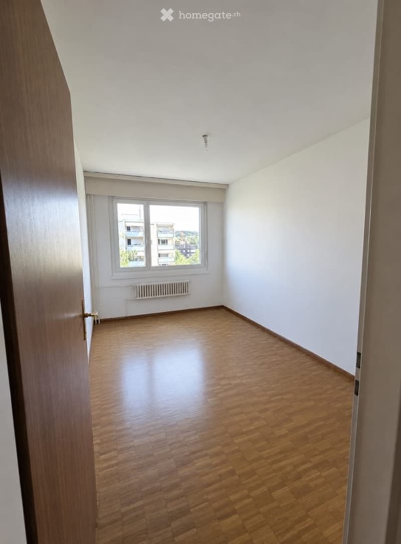 Empty room with wooden flooring, white walls, and a single window with a radiator below it. The room is unfurnished and has a door on the left side.