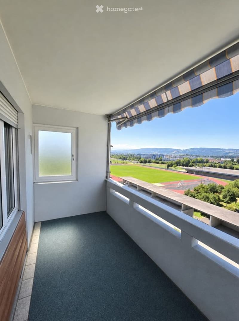 Balcony with a view of a sports field and distant mountains, featuring a striped awning, white walls, and a grey carpeted floor.