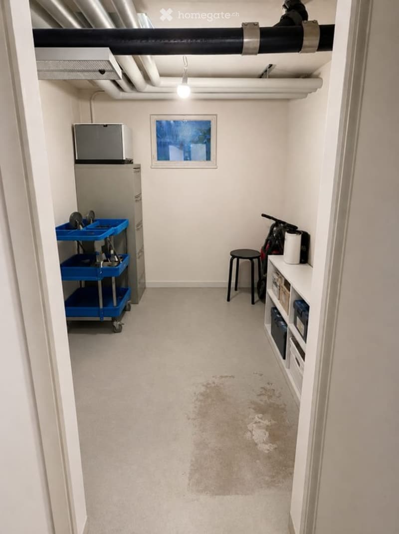 The room is a small, well-lit space with a concrete floor and white walls. It contains a blue rolling cart with tools, a refrigerator, a small stool, shelves with various items, and a bicycle leaning against the wall. There is a window at the far end allo