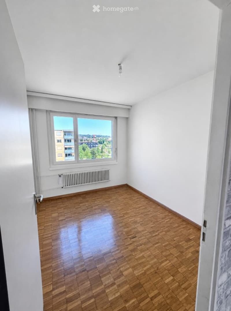 Empty room with wooden flooring, white walls, and a large window offering a city view.