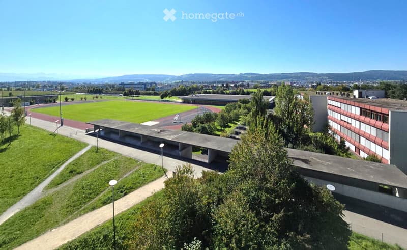 Aerial view of a modern institutional building with a large open green space, pathways, and surrounding trees.