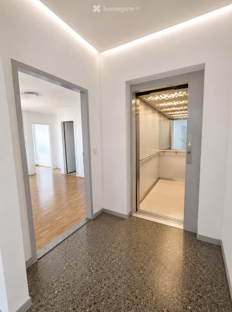 Modern hallway with sleek, contemporary design featuring a polished concrete floor, white walls, and recessed lighting. An elevator is visible through an open doorway, and a mirrored wall reflects the hallway and adjacent rooms.