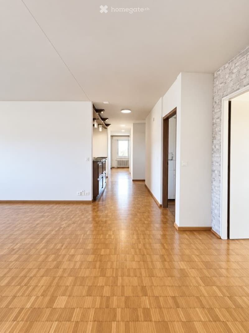 Empty hallway with wooden flooring, white walls, and a visible kitchen area in the background.