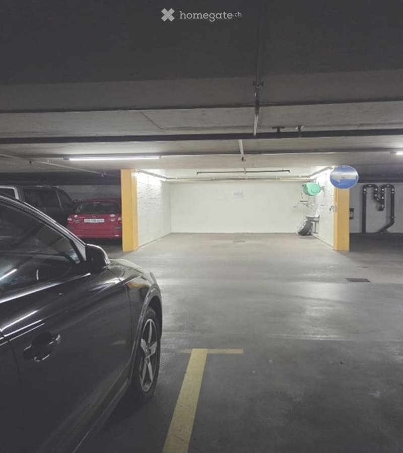 Underground parking area with several parked cars, concrete floors, white walls, and yellow parking lines.