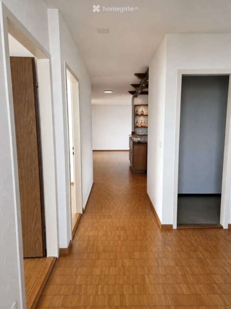 A modern, well-lit hallway with light wooden flooring and white walls. The corridor features several doors and built-in shelving units with decorative items.