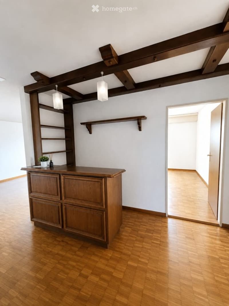 Bright room with wooden flooring and exposed wooden beams on the ceiling. Features a wooden cabinet with drawers, a built-in shelf unit, and pendant lights.