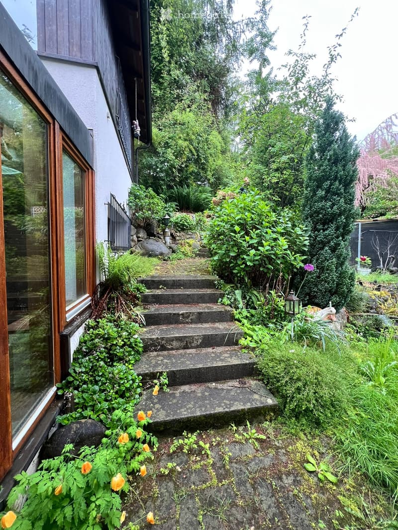 Garden area with stone steps leading up to a house, surrounded by lush greenery and flowers.