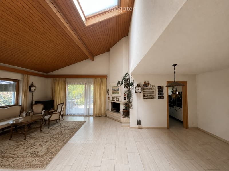 Spacious living room with wooden ceiling, large windows, fireplace, sofa, coffee table, chairs, wall clock, and television.