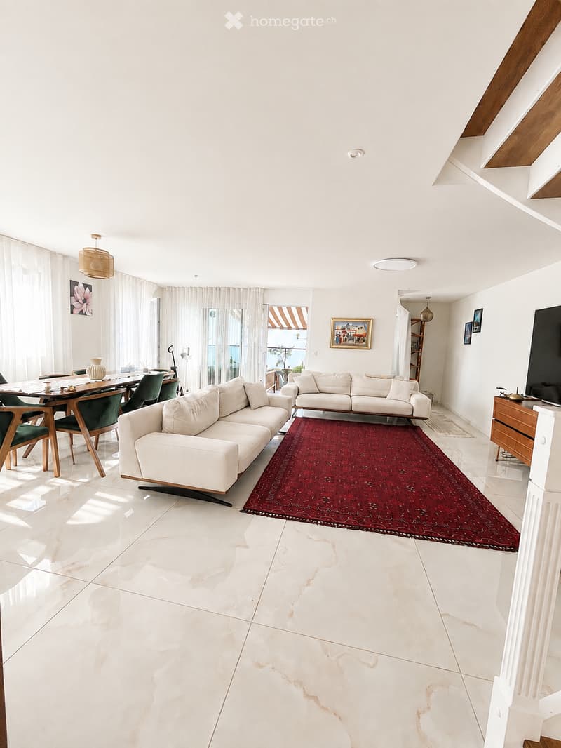 Bright living room with white walls, marble flooring, and large windows. Features a white sectional sofa, dining table with chairs, a red area rug, and decorative items including a vase and picture frames.