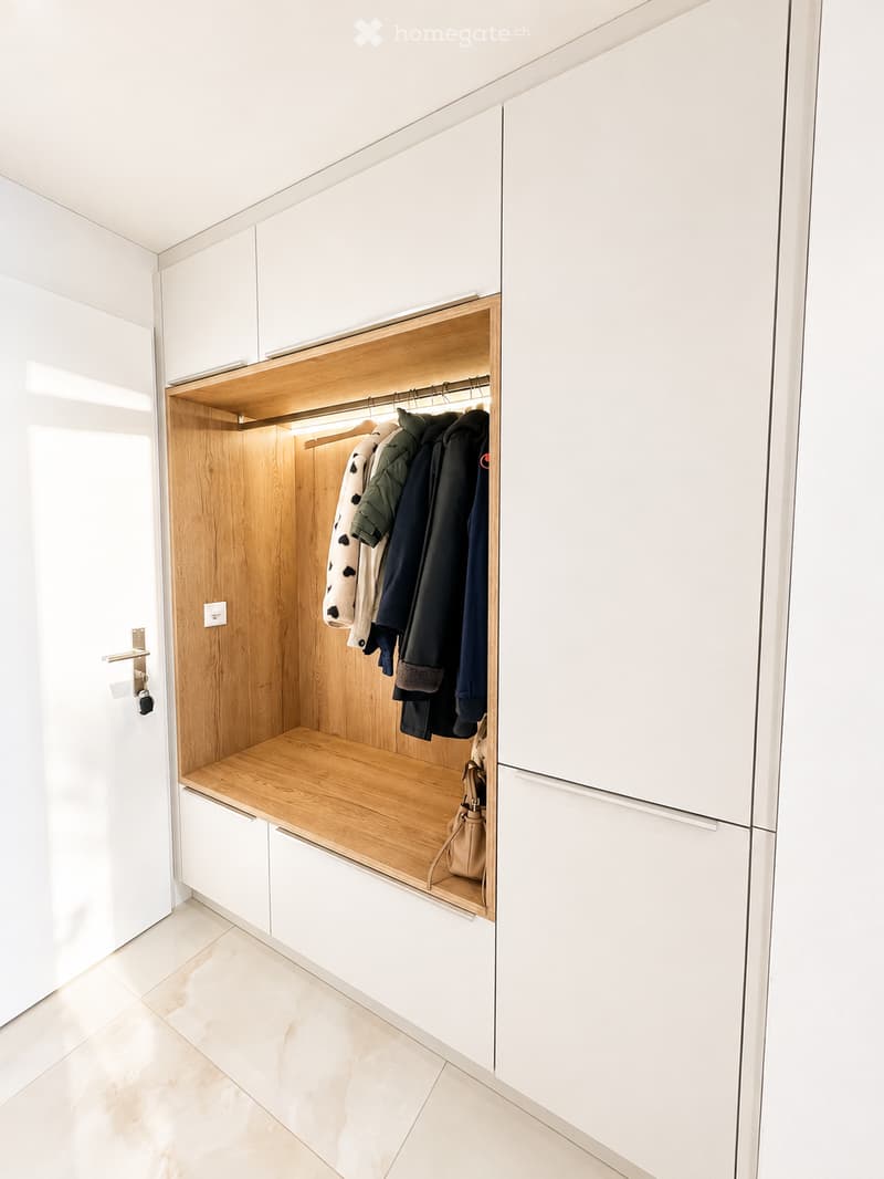 Modern, minimalist hallway with white cabinetry, wooden shelving, and built-in storage. Features a coat rack with jackets and a small bench with a handbag.