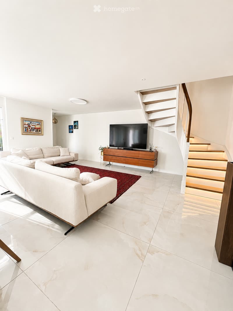 Modern living room with white walls, marble flooring, a large sectional sofa, a coffee table, a TV on a wooden stand, and a staircase leading to an upper level.