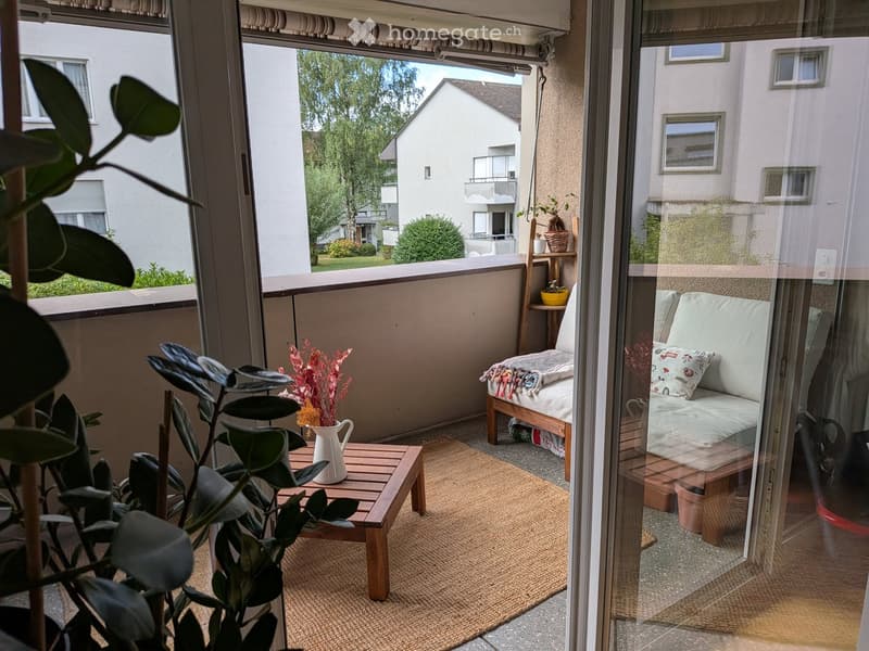 Balcony with a small wooden table, vase with dried flowers, a sofa with cushions, and a rug.