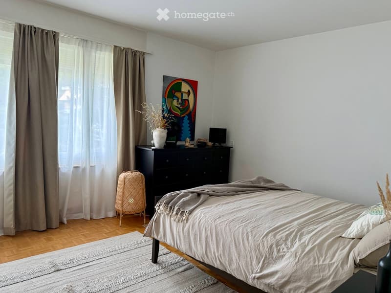 A modern bedroom with a large bed, nightstand, chair, and artwork on the wall. The room features wooden flooring, large windows with curtains, and a cozy, minimalist design.