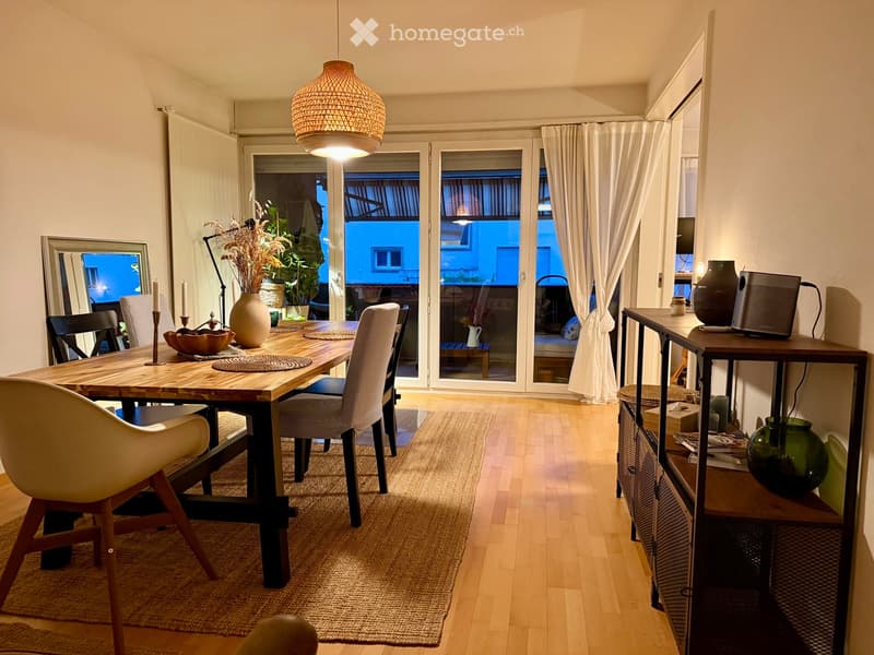 Dining area with a wooden table, chairs, vase, candle, and shelf with decorative items. Large windows with white curtains lead to a balcony.