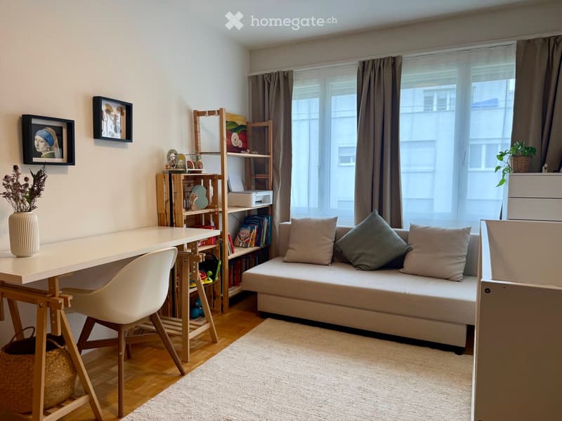 A modern, multi-functional room with a sofa, desk, and shelving unit. The space features a large window with curtains, a wooden floor, and decorative elements like a vase and picture frames.
