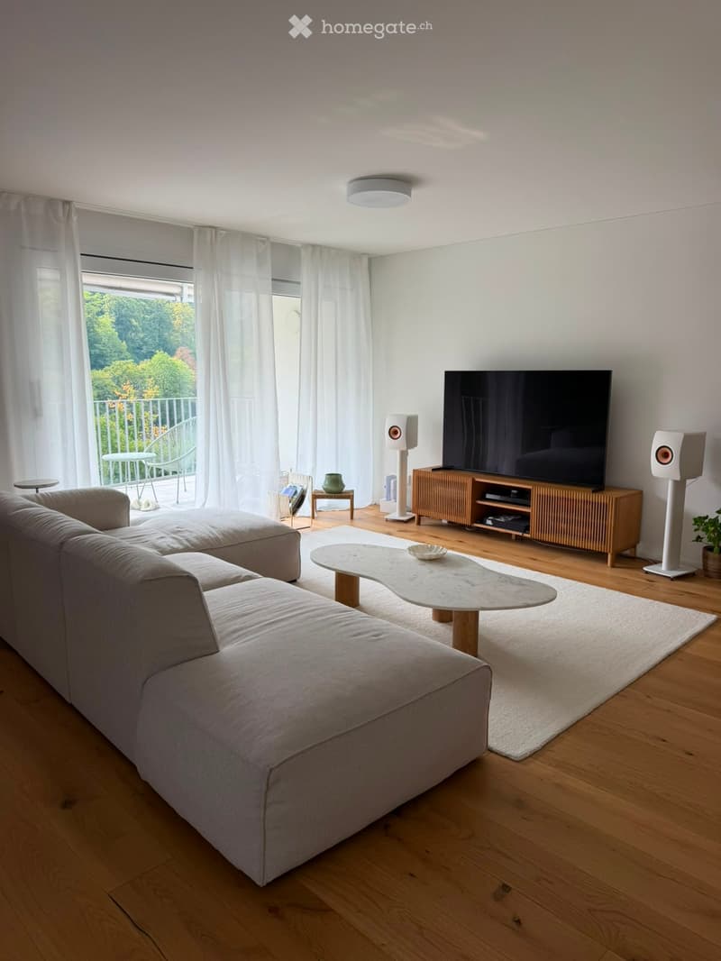 Modern living room with a grey sectional sofa, a white coffee table, a large flat-screen TV on a wooden stand, and two white speakers. The room features wooden flooring, large windows with white curtains, and a balcony with outdoor furniture visible throu