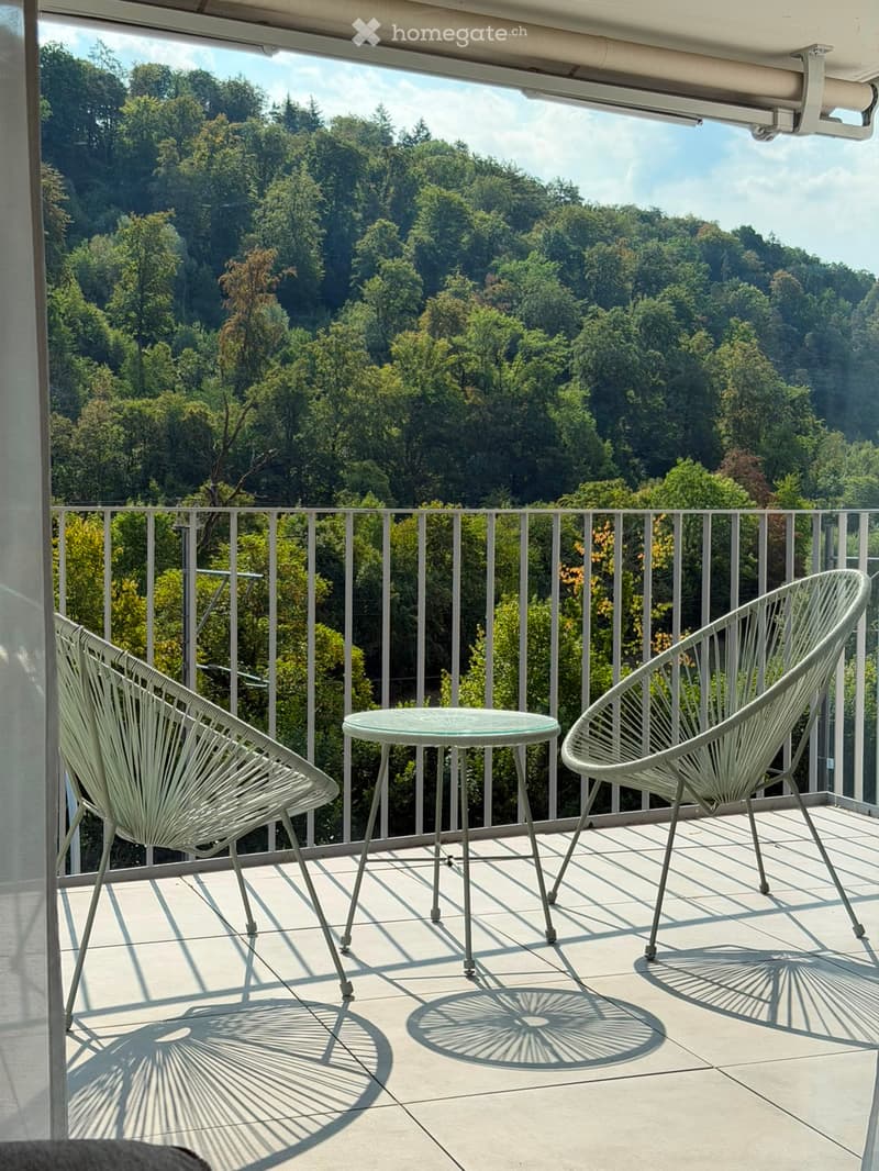 Balcony with a scenic view of a forested area, featuring two white wicker chairs and a small round table.