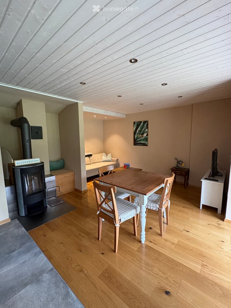 Open-plan living and dining area with wooden flooring, a fireplace, a sofa, a dining table with chairs, and a TV screen.