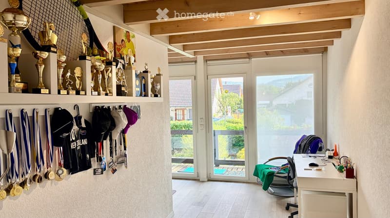 A well-lit room with a wooden ceiling, trophies and medals on a shelf, sports ribbons and hats hanging on the wall, a desk with a chair, and a large window with a view of the garden.