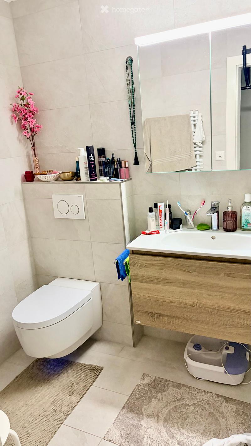 Modern bathroom with a toilet, sink, mirror, and various toiletries on the counter. Features a wooden vanity, a glass shower enclosure, and decorative elements like a vase with pink flowers and a small rug.
