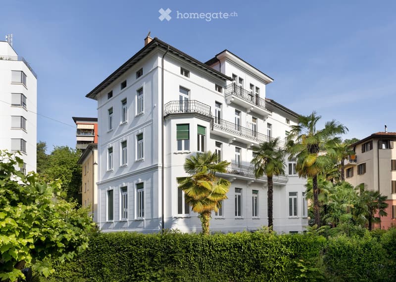A large, white multi-story building with multiple windows and balconies, surrounded by greenery and palm trees.