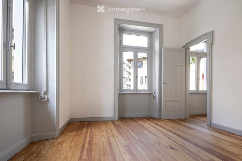 Empty room with light grey walls, wooden flooring, and multiple windows and doors leading to other spaces.