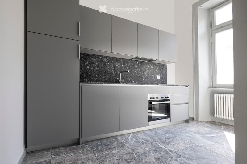 Modern kitchen with grey cabinets, marble backsplash, and a built-in oven. Features a tiled floor and a window providing natural light.