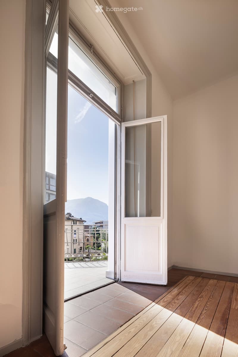An unfurnished attic room with wooden flooring, large windows, and a view of a cityscape and mountains through an open door leading to a tiled balcony.