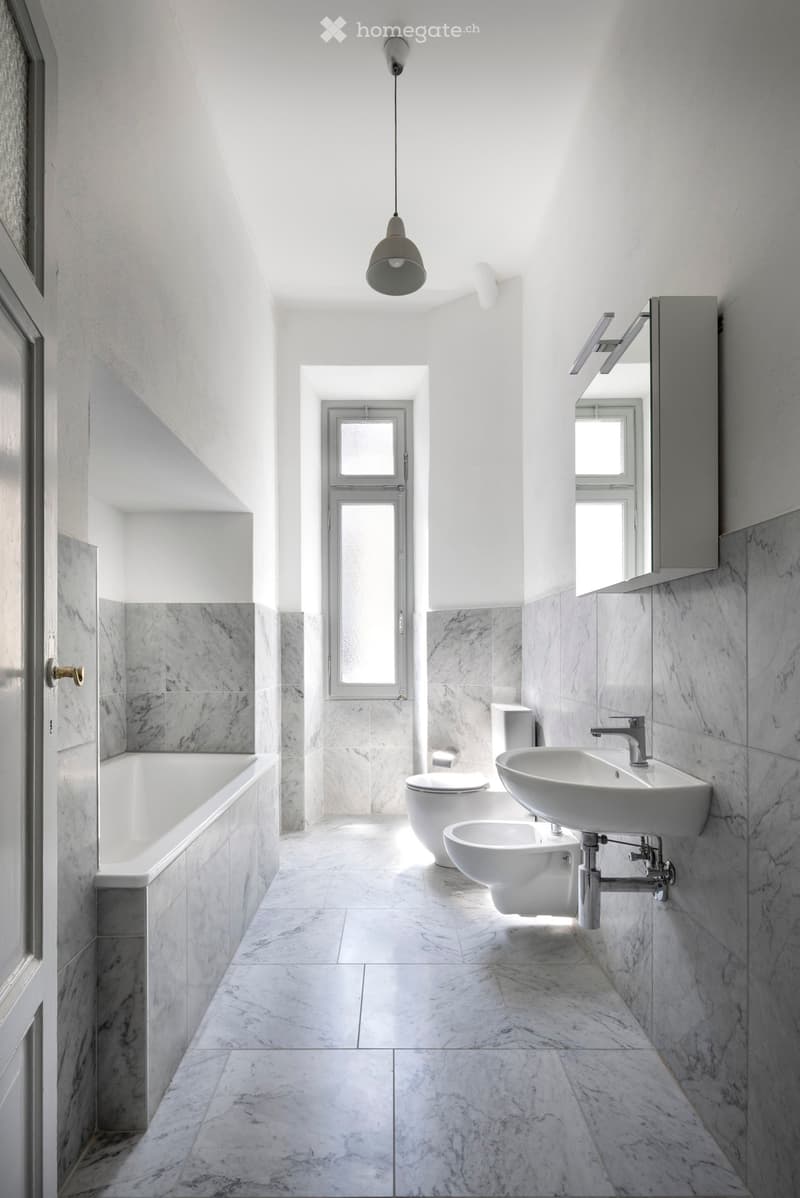 Modern bathroom with marble tiles, white fixtures, and natural light from windows.