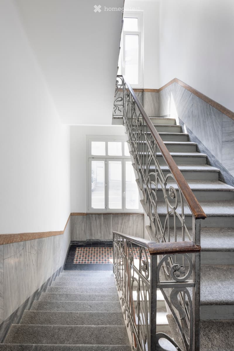 Staircase with grey steps, wooden handrail, metal balustrade, and patterned tile landing area. Natural light from windows illuminates the space.