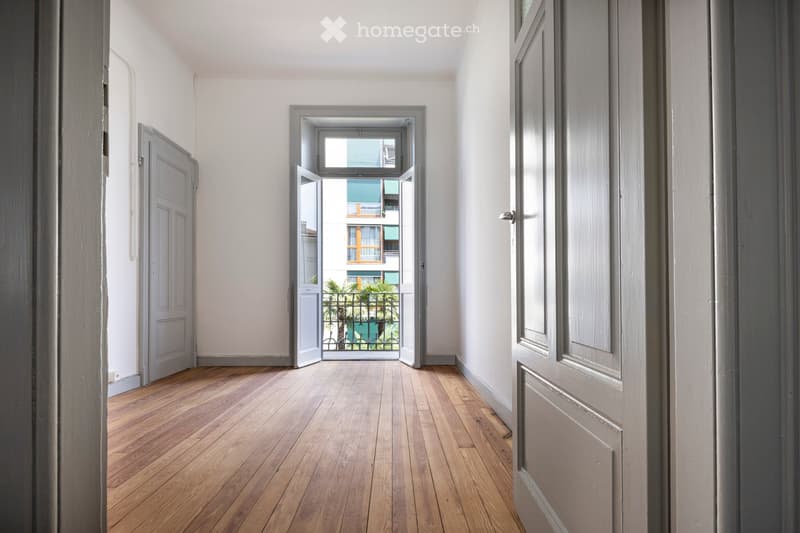 Empty hallway with wooden flooring, white walls, and doors on both sides. An open doorway leads to a balcony with a view of a building and greenery.