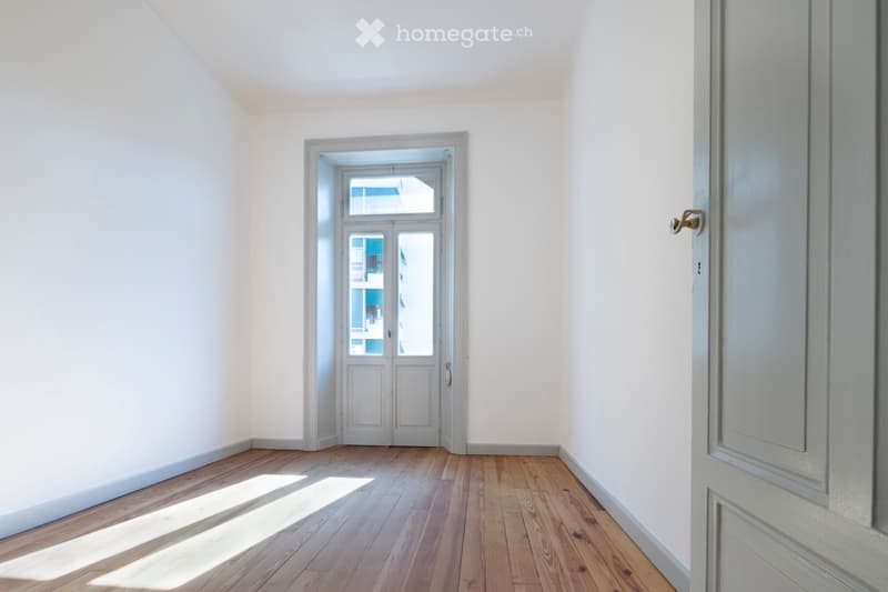 Empty room with light wooden flooring and white walls, featuring a doorway leading to another room with a glass door and visible outdoor view.