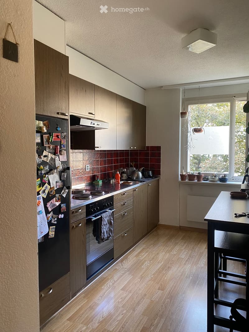 Modern kitchen with light wood cabinets, stainless steel appliances, and a central island. Features a refrigerator with magnets, a stove, oven, and a dining area with a table and chairs.