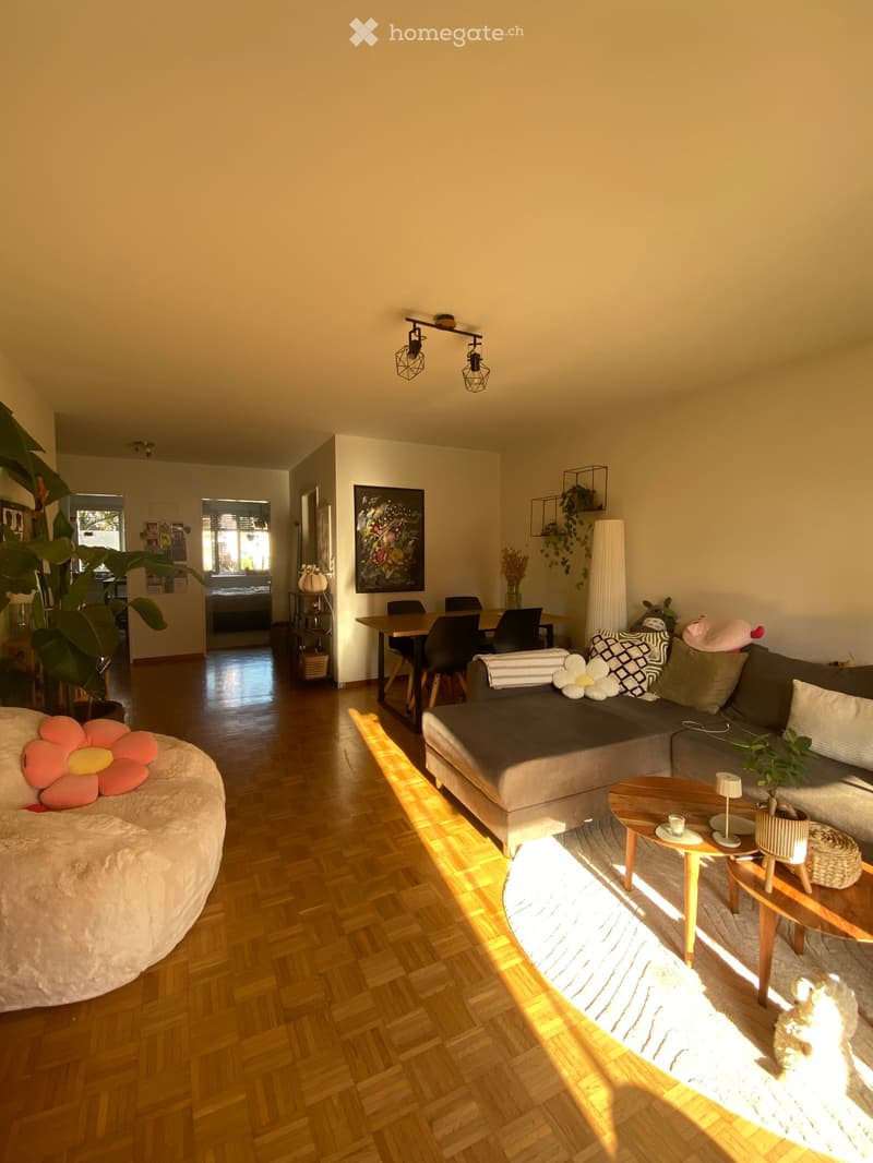 Bright living room with wooden flooring, a grey sectional sofa, a round coffee table, a dining table with chairs, and decorative plants.