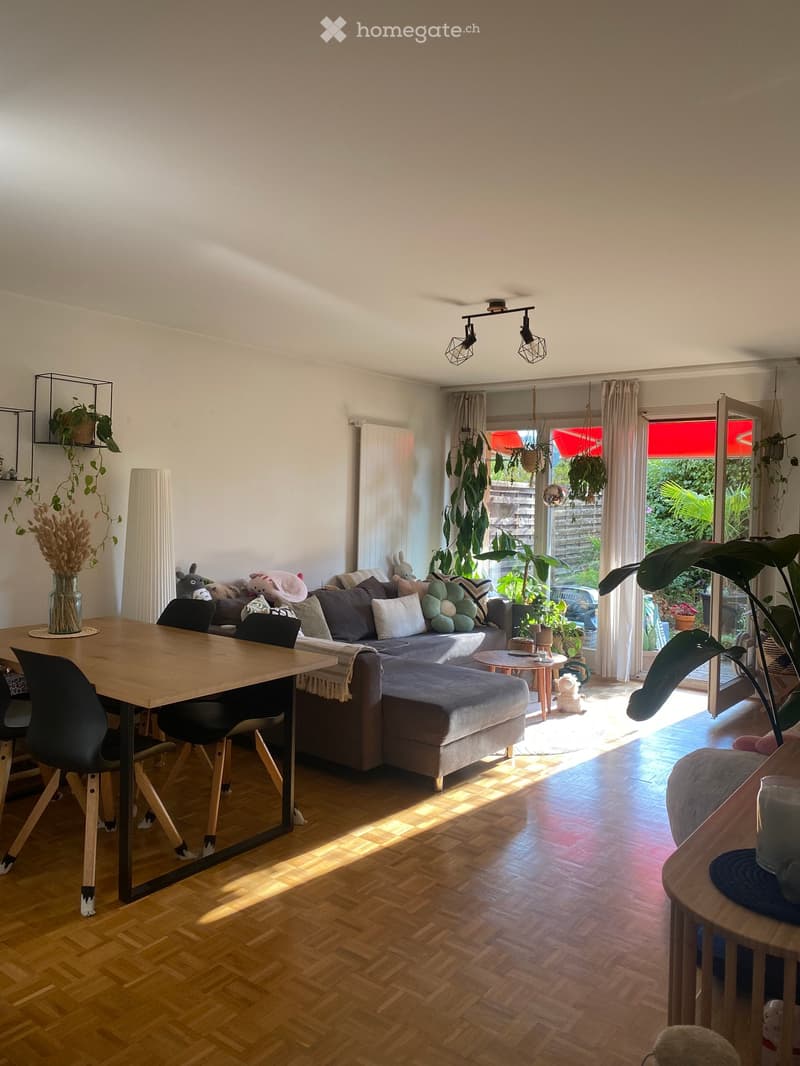 Bright living room with wooden flooring, large window with sheer curtains, and a sliding glass door leading to a garden. Features a grey sectional sofa with multiple cushions, a wooden coffee table, a dining table with chairs, and several potted plants.